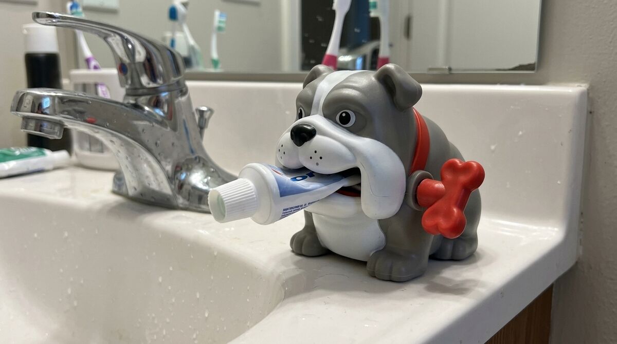 A gray and white bulldog squeezer with a red collar and bone, gripping a toothpaste tube